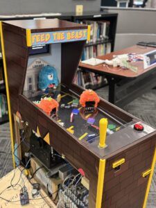 A homemade arcade game titled "FEED THE BEAST," built for the Engineering Capstone Showcase by Lakota East and Butler Tech students, features fuzzy monster figures, electronics, a yellow joystick, and sits against a backdrop of library bookshelves.