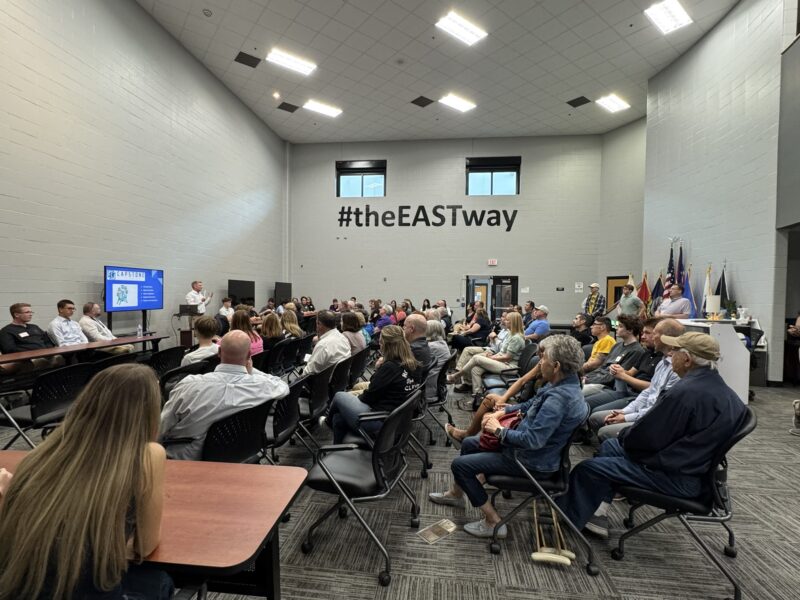 A group of people sit in rows facing a panel and a presentation screen in a large, modern room with high ceilings. The phrase