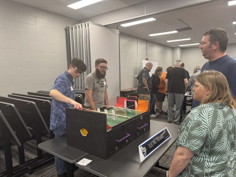Two young men from Lakota East demonstrate a pinball machine on a table to a group at the Butler Tech Engineering Capstone Showcase. Other attendees are visible in the brightly lit room with gray walls.