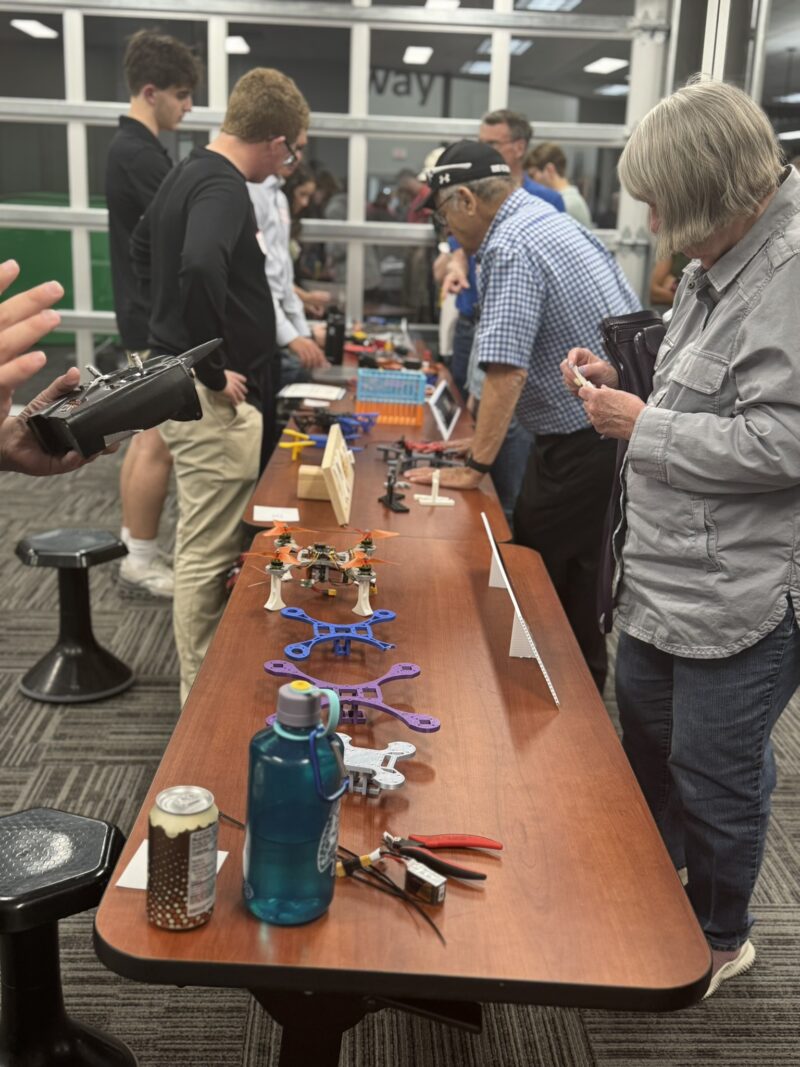 People gather around a table at the Engineering Capstone Showcase, displaying small drones, tools, and water bottles. Some examine items closely while others talk or observe in the bright, carpeted room at Lakota East Butler Tech.