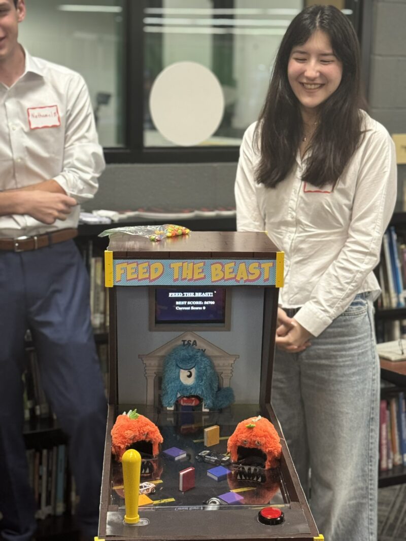 A young woman stands smiling beside a tabletop arcade game called