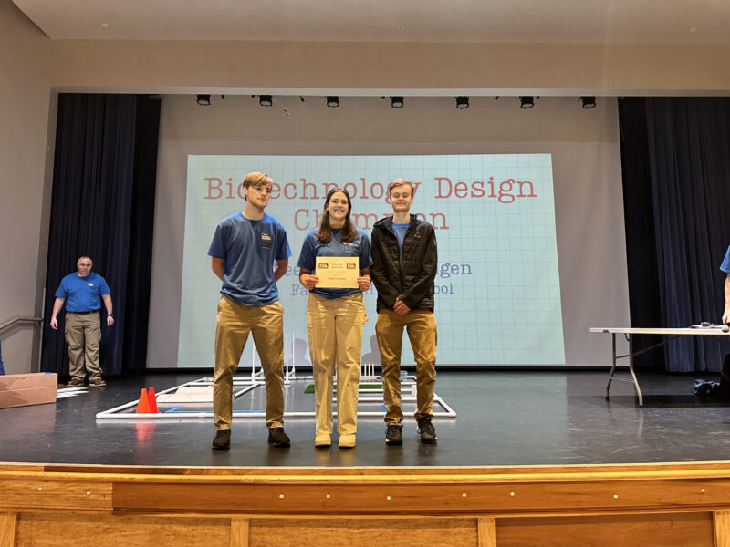 Three students from Butler Tech stand onstage holding a certificate at a Biotechnology Design Challenge event. A screen behind them displays the event name, while two adults and STEM-themed materials are visible on stage.