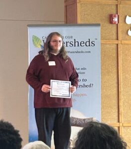 Gavin Owens, dressed in a maroon sweater and dark pants, stands on stage holding a certificate for Environmental Innovation, as several audience members watch the presentation.