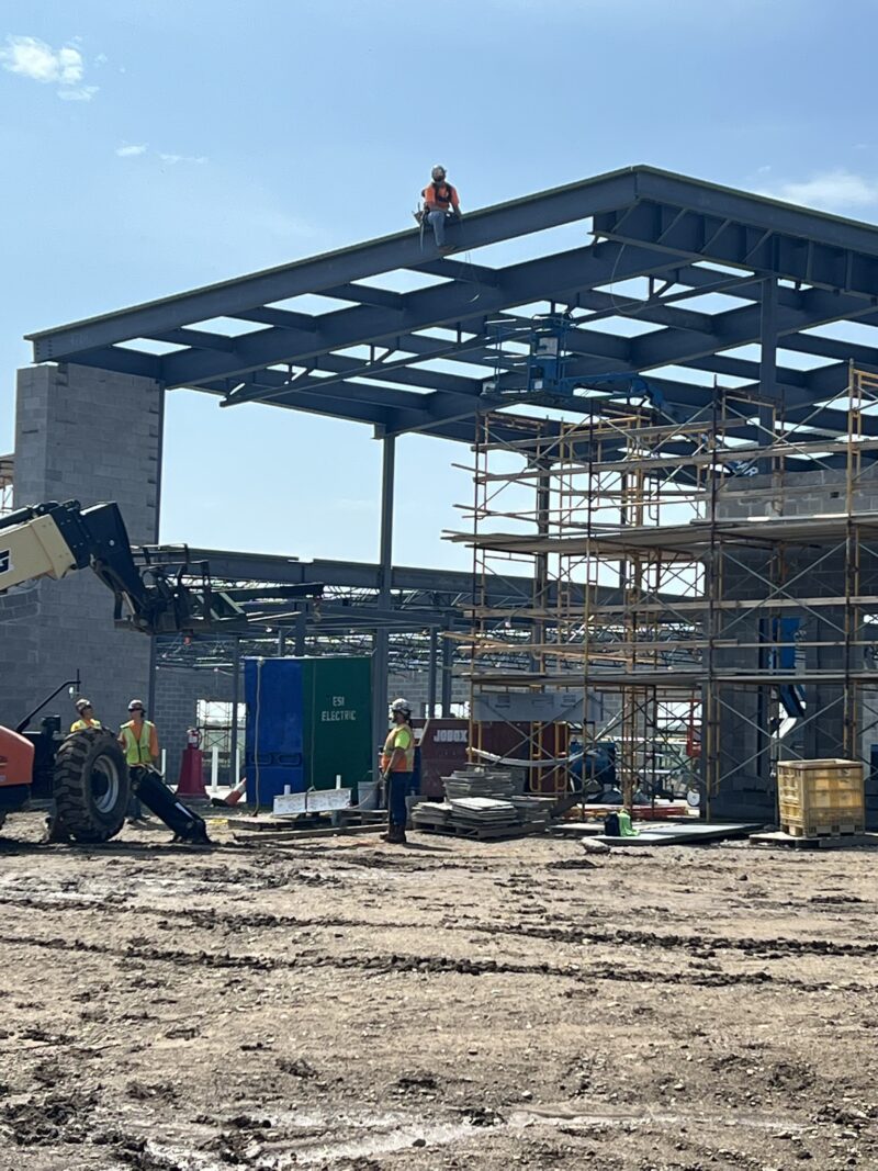 Construction workers in safety vests and helmets navigate scaffolding and equipment at the Butler Tech Aviation Center, while one worker sits atop the steel framework, marking a topping out milestone under a clear sky.