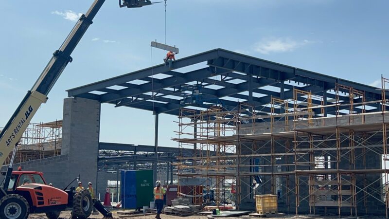 Construction workers at Butler Tech’s Aviation Center use cranes and scaffolding to build a large steel-framed structure under a clear sky, celebrating the Topping Out Milestone as one worker is elevated on the roof while others work below.