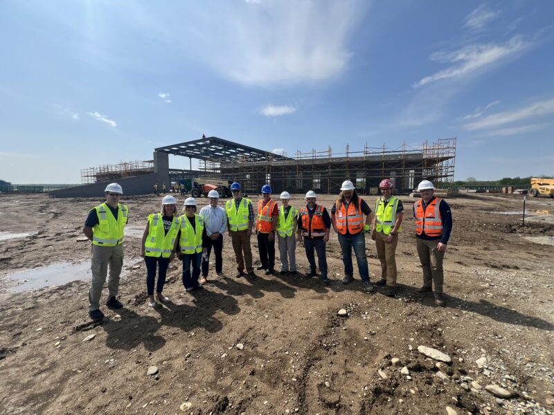 A group of people wearing safety vests and hard hats stand on the Butler Tech Aviation Center construction site, celebrating a Topping Out Milestone with a partially built steel structure and scaffolding under a partly cloudy sky.