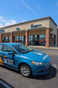 A blue Butler Tech driver education car is parked in front of a Butler Tech building in Milford on a sunny day. The building features large windows and brick accents.