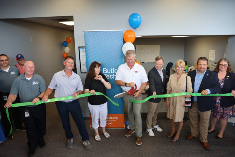 A group of people stands in an office, smiling as a man in the center cuts a green ribbon with large scissors, celebrating a Butler Tech Driver Education event. Blue and orange balloons and a Butler Tech banner are in the background.