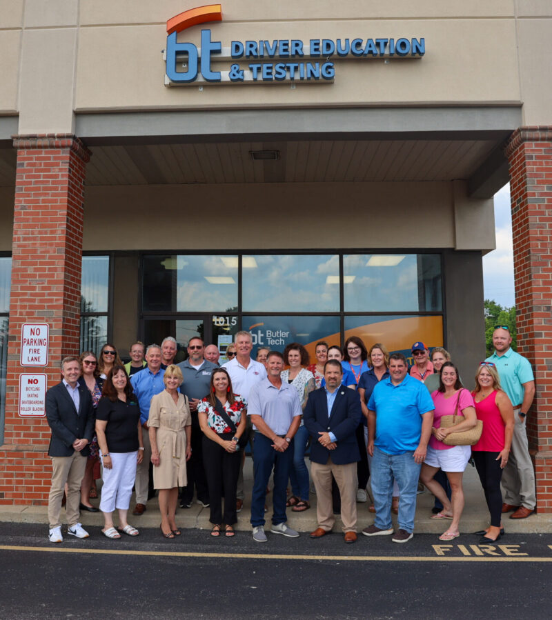 A large group of people pose and smile together outside the Milford driver education & testing site on a sunny day. The group stands near the curb, with parked cars and trees in the background.