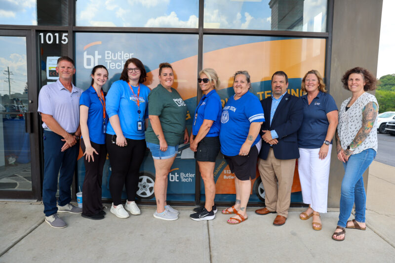 Nine adults stand in a row, smiling, outside a building with glass windows displaying the Butler Tech logo—home to Milford's premier Driver Education and Testing Site. Most wear blue shirts and casual attire; two are dressed in business attire.