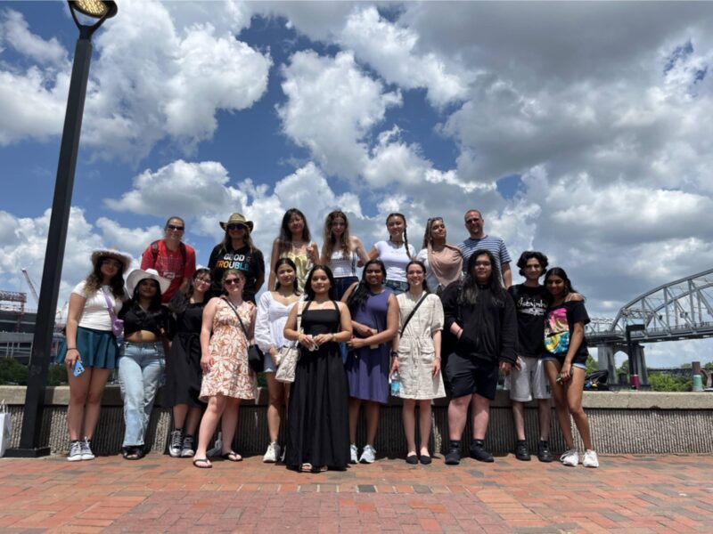 A group of seventeen people pose together outdoors on a sunny day, standing on a brick patio with the Nashville cityscape and a bridge in the background, representing Butler Tech at HOSA International.