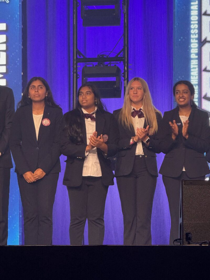 Four young women in dark suits and bow ties stand on stage at HOSA International in Nashville, clapping and facing forward, with a blue-lit background and stage lighting behind them.