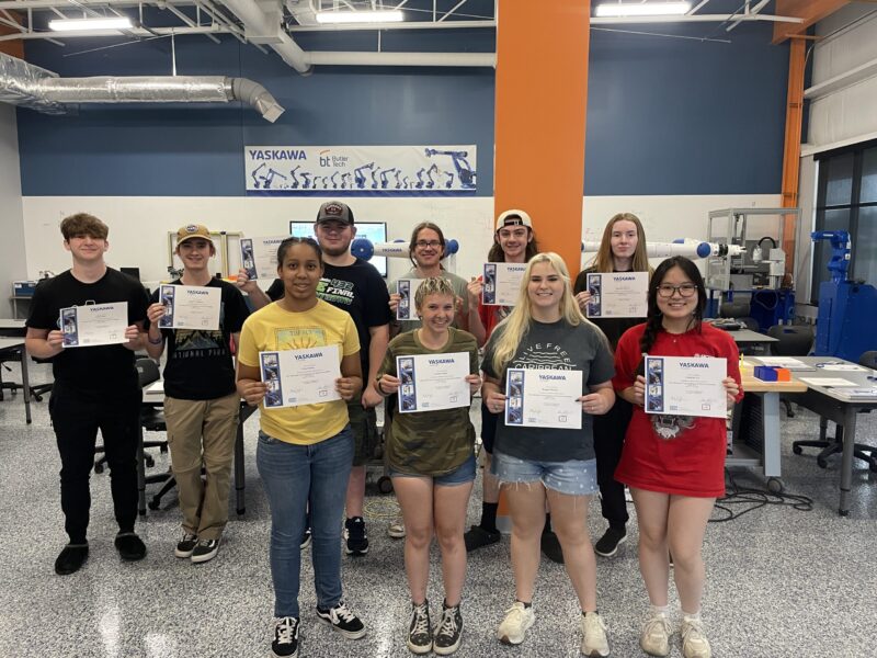 A group of ten high school students stand indoors, smiling and holding certificates, with industrial equipment and a “Yaskawa” sign in the background, celebrating their Yaskawa Credential earned through a summer training program.