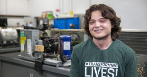 A young man with wavy hair wearing a "#TRANSFORMING LIVES!" t-shirt smiles while sitting in a Butler Tech workshop or lab, surrounded by technical equipment and tools, preparing for in-demand careers.