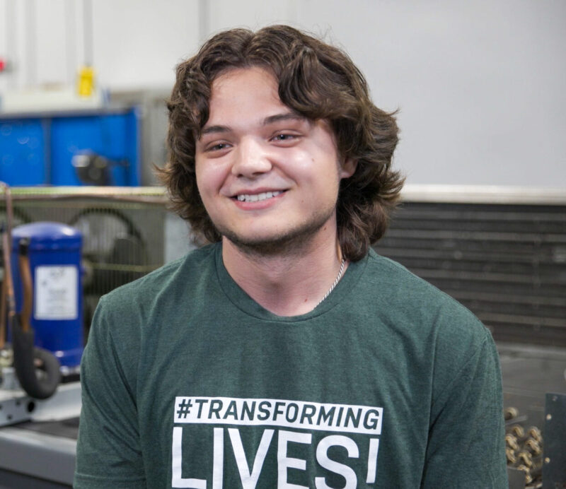 A young person with wavy hair smiles while wearing a green “#TRANSFORMING LIVES!” T-shirt, sitting in a Butler Tech workshop filled with technical equipment and tools, preparing for in-demand careers.