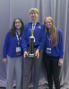 Three smiling people wearing blue shirts and conference badges stand together in front of a gray curtain at the TSA Conference, with the person in the center holding a gold trophy.