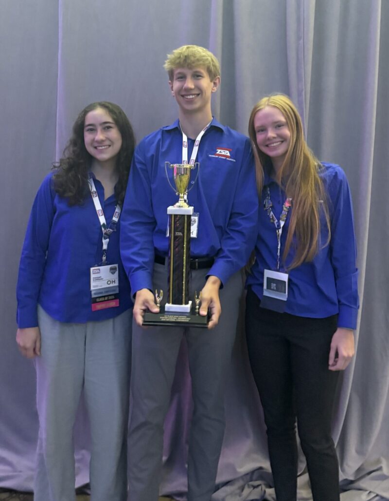 Three smiling people wearing blue shirts and conference badges stand together in front of a gray curtain at the TSA Conference, with the person in the center holding a gold trophy.