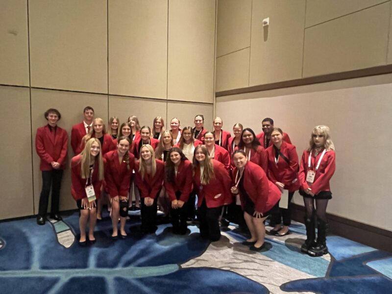 A group of young adults from Butler Tech, wearing red blazers and name badges, pose together in a conference room. Smiling FCCLA Finalists, some stand while others crouch in front against beige walls and a blue-patterned carpet.