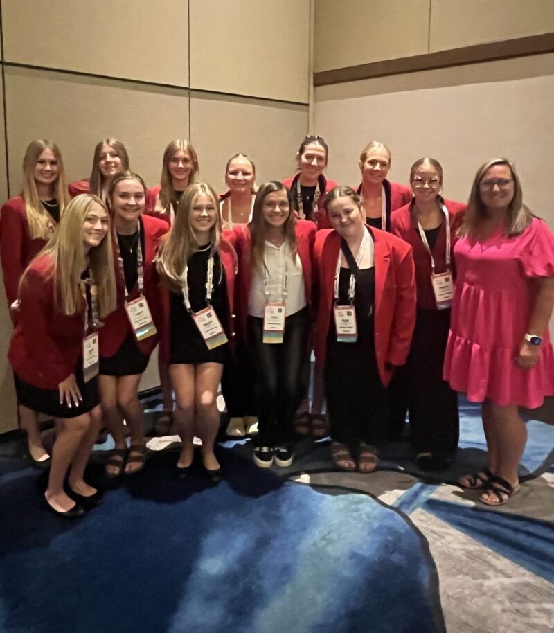 A group of thirteen women, including FCCLA Champions and National Finalists, pose together indoors in red blazers and black outfits, while one on the right stands out in a bright pink dress. They smile on a blue and gray patterned carpet.