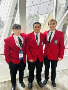 Three young men stand indoors, smiling at the camera in matching red blazers with SkillsUSA Championships patches. One wears a conference badge. Large windows reveal a cityscape, highlighting their national recognition with Butler Tech.