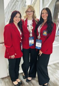 Three young women from Butler Tech stand indoors in matching red blazers, black pants, and white shirts. Wearing name badges and medals, they smile proudly after earning national recognition at the SkillsUSA Championships. Large windows reveal a city view behind them.