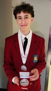 A young person from Butler Tech wearing a red blazer, white shirt, and black tie smiles while holding a SkillsUSA champion medal and case; a white medal ribbon is around their neck, celebrating national recognition at the SkillsUSA Championships.