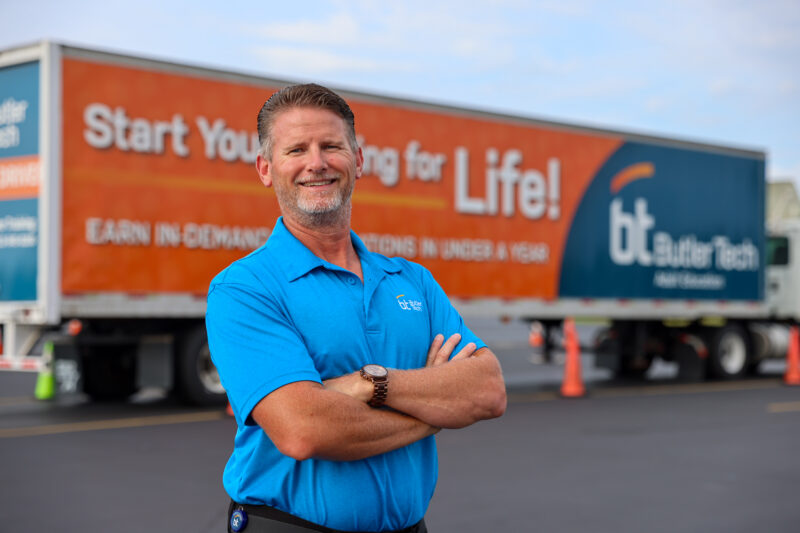 A man in a bright blue polo stands smiling with arms crossed in front of a large truck with an orange and blue trailer that reads, 