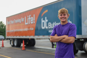 A young man in a purple shirt stands smiling with arms crossed in front of a large truck trailer with "Butler Tech" branding, orange CDL Training signage about training for life, and orange traffic cones nearby.