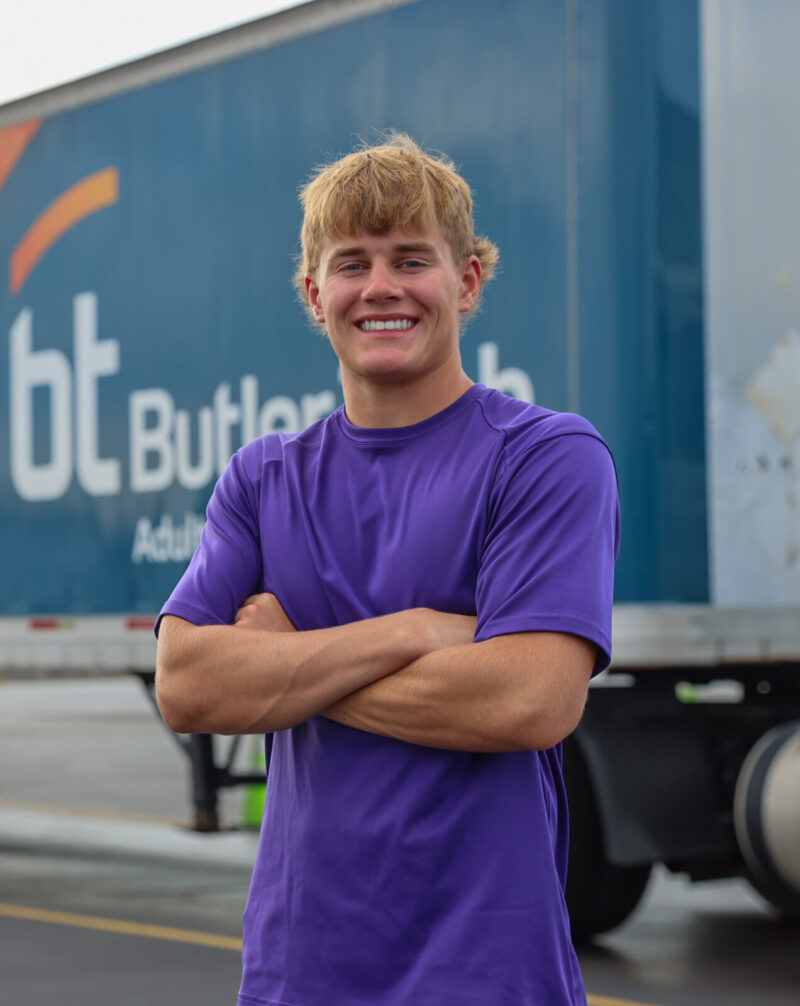 A young man in a purple shirt stands smiling with arms crossed in front of a large semi trailer that reads “Start Your Training for Life!” and “bt Butler.” Orange traffic cones line the pavement, highlighting the importance of CDL driver training.