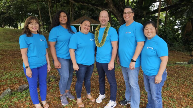 Six people stand outdoors on grass and fallen leaves, wearing matching bright blue shirts. One man in the center wears a leafy lei. Trees and greenery fill the background. The group is smiling and facing the camera.