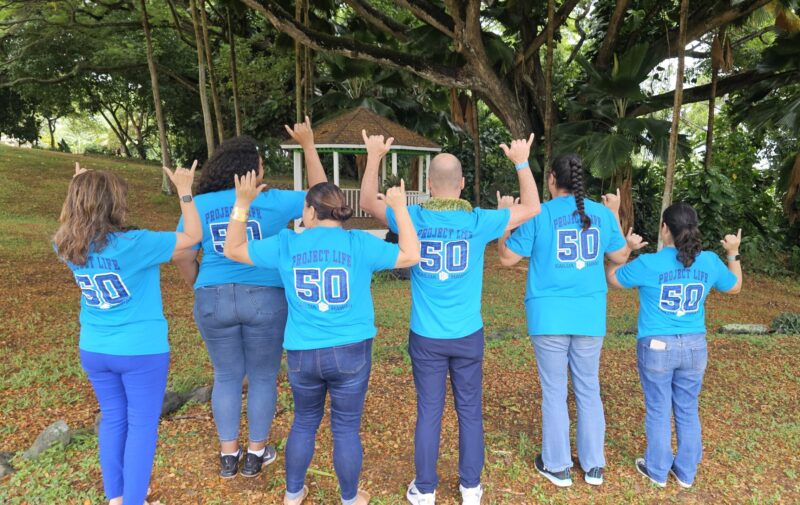 Six people stand outdoors with their backs to the camera, wearing matching bright blue shirts with