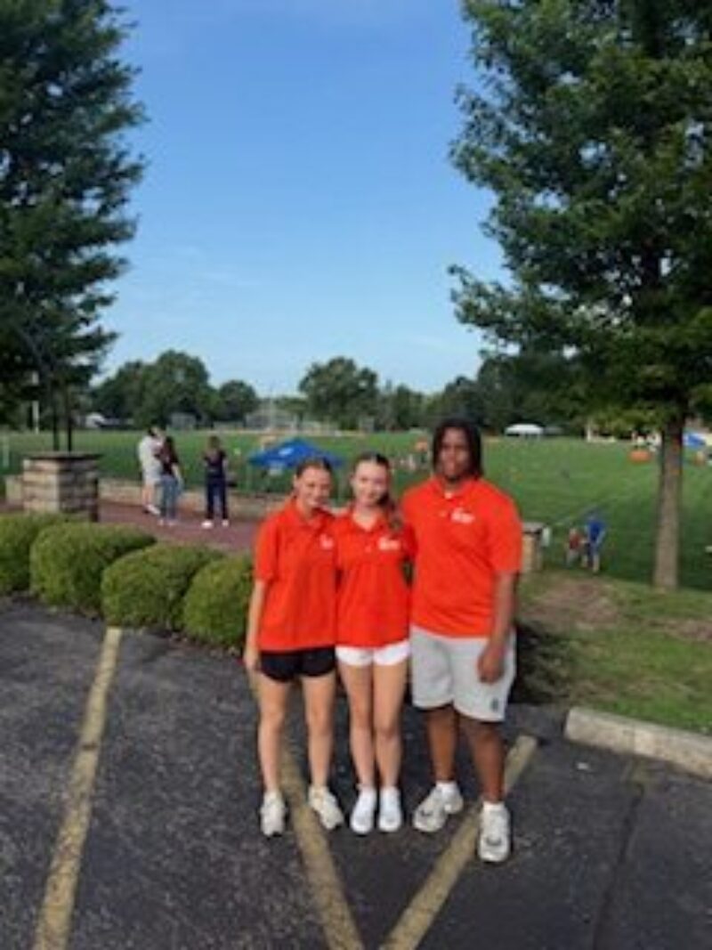 Three Exercise Science students wearing matching orange shirts and shorts stand together smiling in a parking lot near a grassy field, with trees and people in the background on a sunny day.