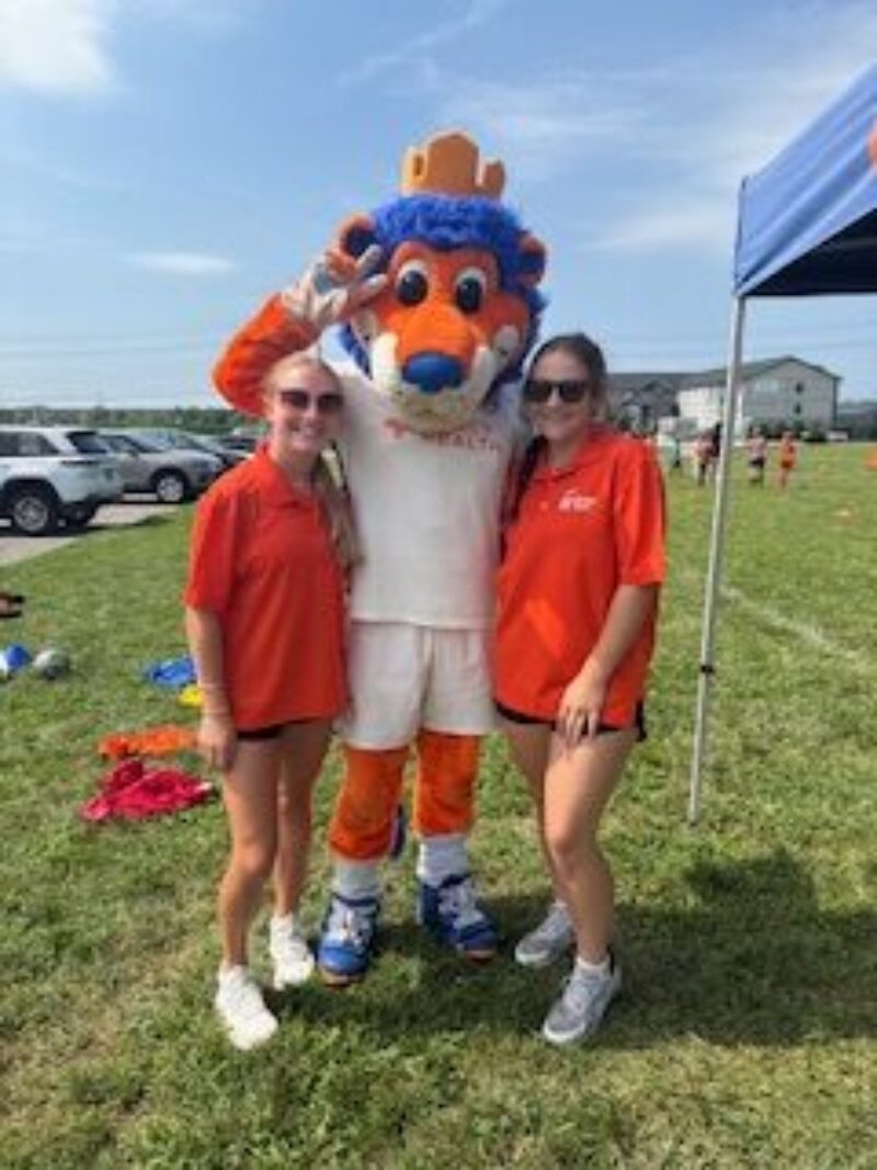 Two Exercise Science students in orange shirts stand on either side of a blue and orange FC Cincinnati mascot in a crown, all smiling outdoors on a grassy field under a sunny sky.