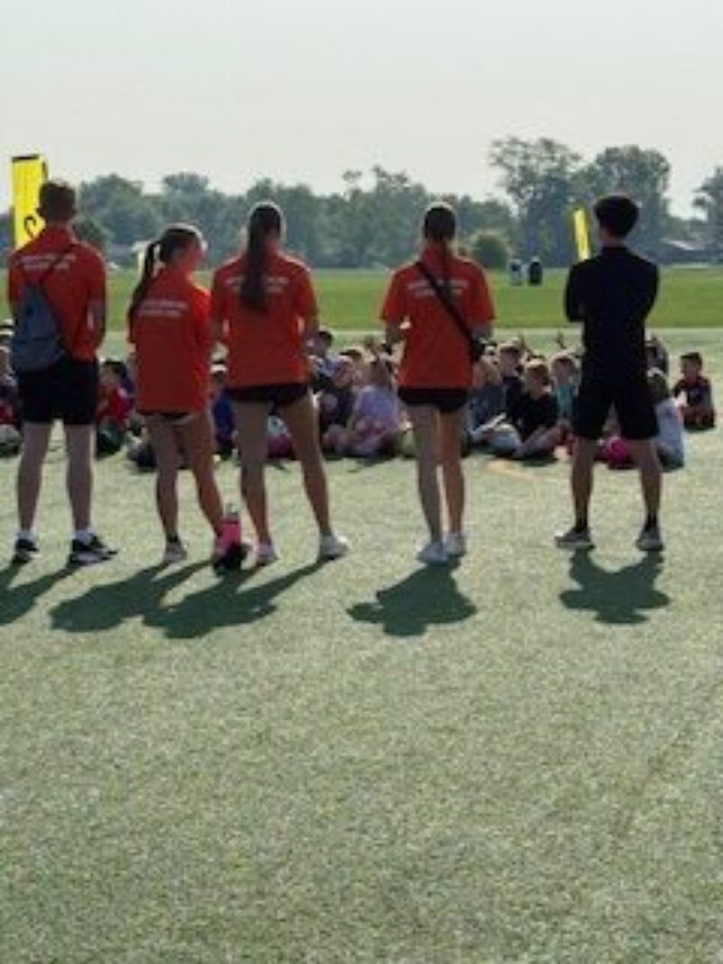 Five students stand in front of a seated crowd on a grassy field. Four wear matching orange shirts, one wears dark clothes. Trees and yellow banners are visible in the background under a clear sky at this FC Cincinnati event.