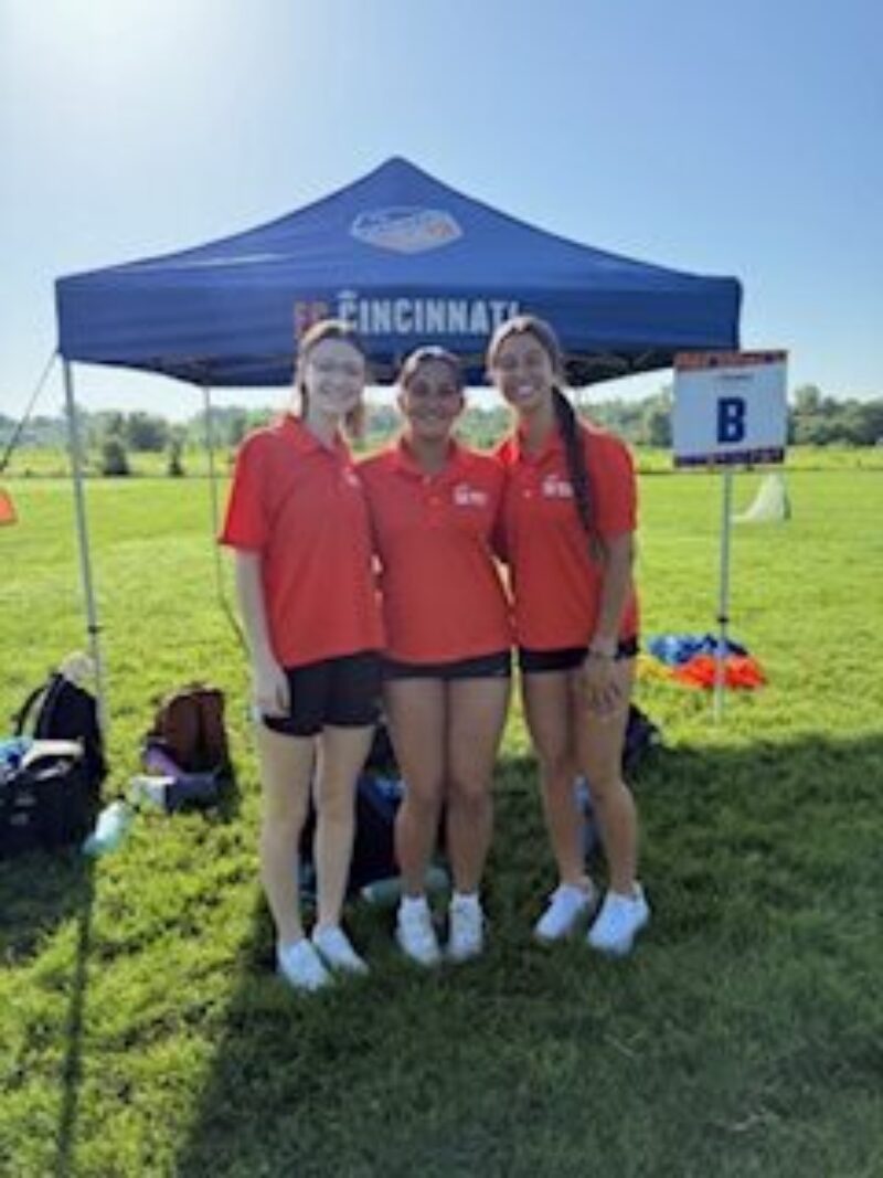 Three young women wearing matching red shirts and shorts stand smiling together in front of a blue tent labeled 