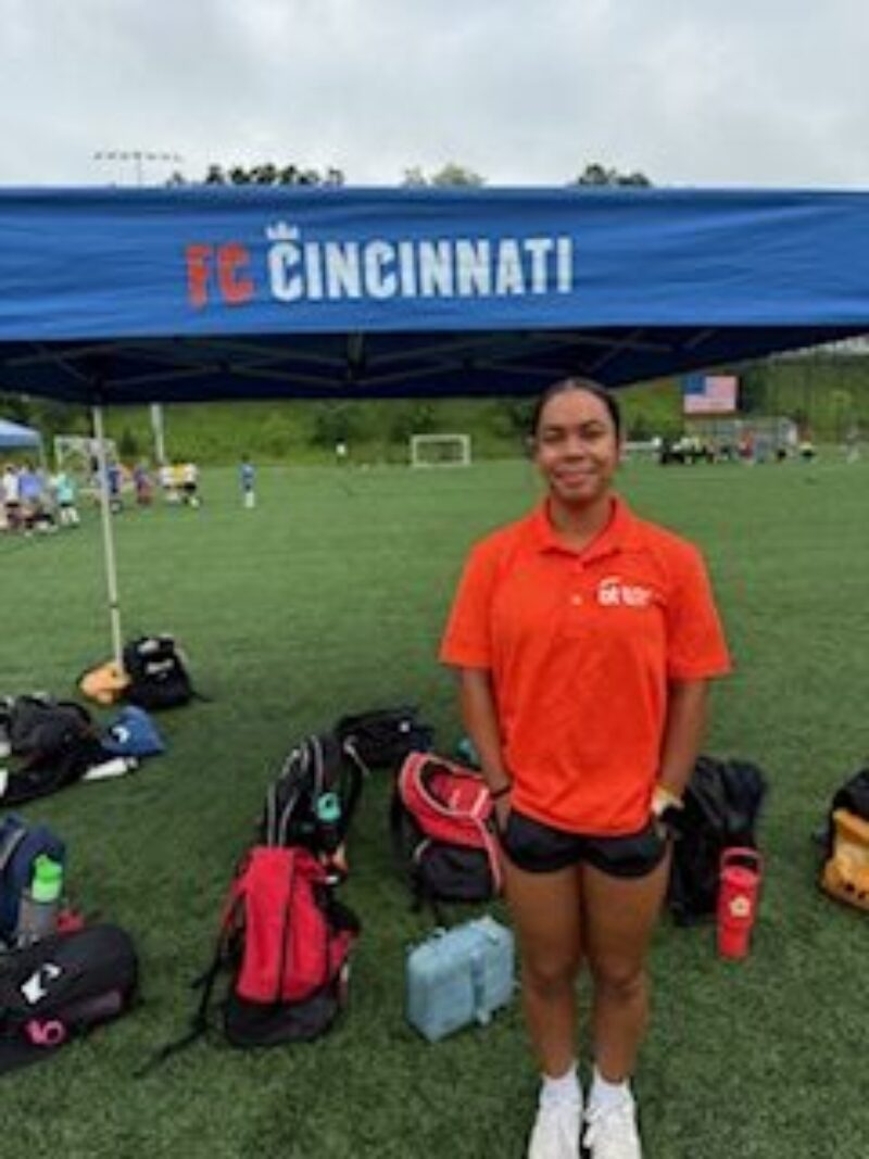 A young woman in an orange FC Cincinnati shirt stands on a soccer field in front of a blue FC Cincinnati tent, surrounded by soccer bags and gear, representing students interested in exercise science and sports.