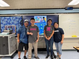 Four people stand smiling in a classroom in front of a screen displaying a "Portrait of a Graduate" graphic. The group poses together, with a whiteboard and blue patterned wall decor in the background.