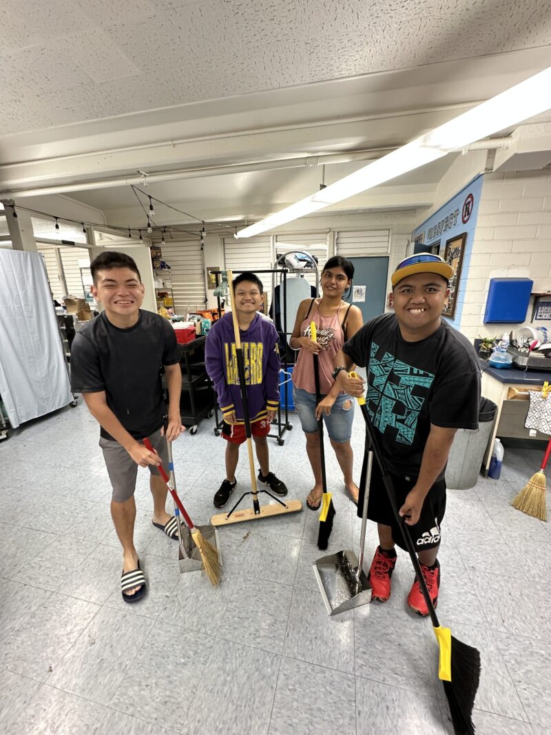Four young people stand indoors holding cleaning tools, such as brooms and dustpans, smiling at the camera. They appear to be part of a cleaning activity in a bright, organized room with supplies in the background.