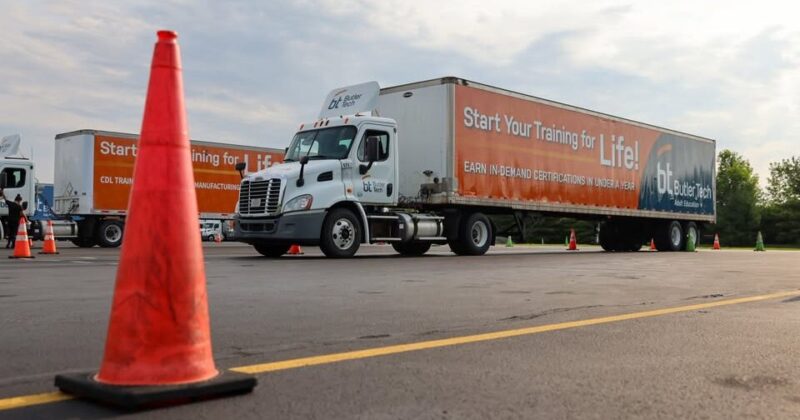 A large orange and white semi-truck with “Start Your Training for Life!” printed on the trailer is parked in a lot, with an orange traffic cone in the foreground and more cones in the distance.