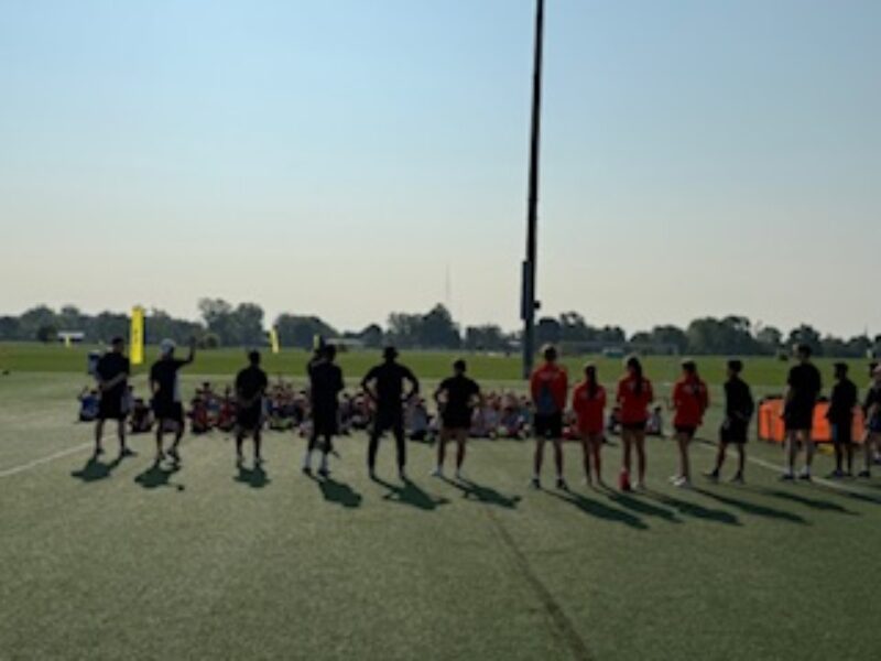 A row of students stand on a grassy sports field with long shadows, facing a group of seated individuals in the background on a sunny day. Trees and sports equipment are visible in the distance.