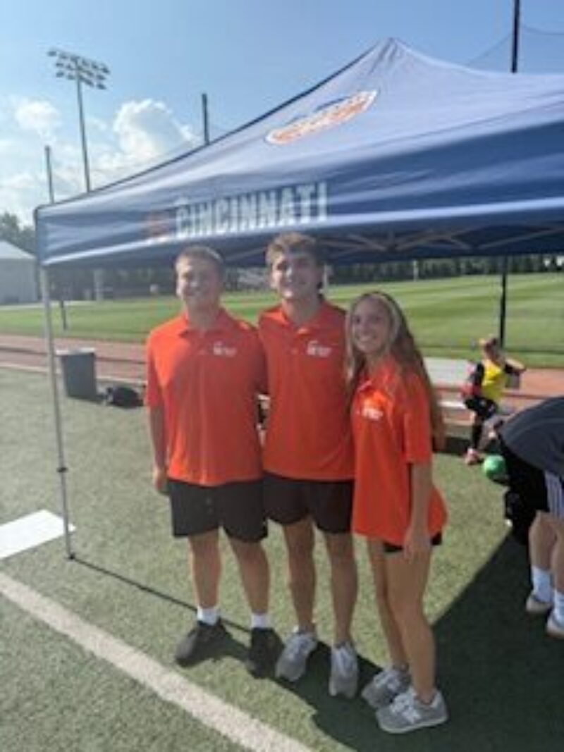 Three students wearing matching orange shirts and black shorts stand and smile in front of a blue tent with 