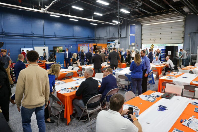 A large group gathers in a spacious, brightly lit room with tables covered in orange and white tablecloths. Some are seated and talking, while others stand and mingle near Butler Tech Business Advisory Council displays and equipment.