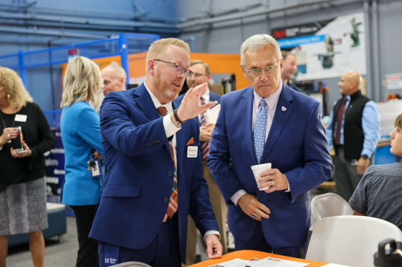 Two men in blue suits talk at an indoor event; one gestures with his hand while the other holds a cup. Several people and industrial equipment, along with Butler Tech Workforce Leadership members, are visible in the busy background.