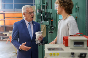 An older man in a blue suit talks to a younger man in a gray t-shirt inside an industrial workshop, with machinery and tools visible—highlighting Butler Tech’s commitment to workforce leadership and hands-on training.