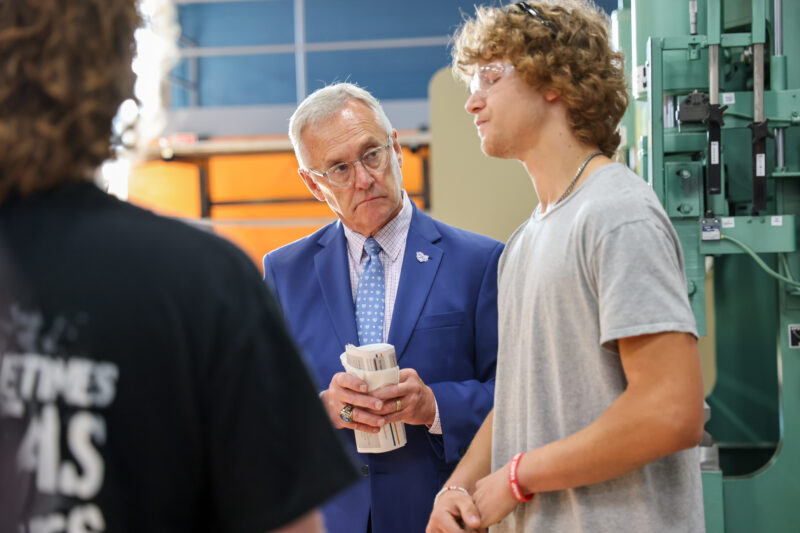 A man in a blue suit and glasses listens attentively to a young man in a gray T-shirt and glasses at Butler Tech, highlighting Workforce Leadership as another individual stands nearby with their back to the camera.