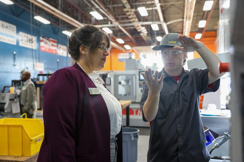 A woman in business attire listens as a man in work clothes and safety glasses explains something, gesturing with his hands, in an industrial workshop—a Workforce Leadership discussion at Butler Tech’s Business Advisory Council.