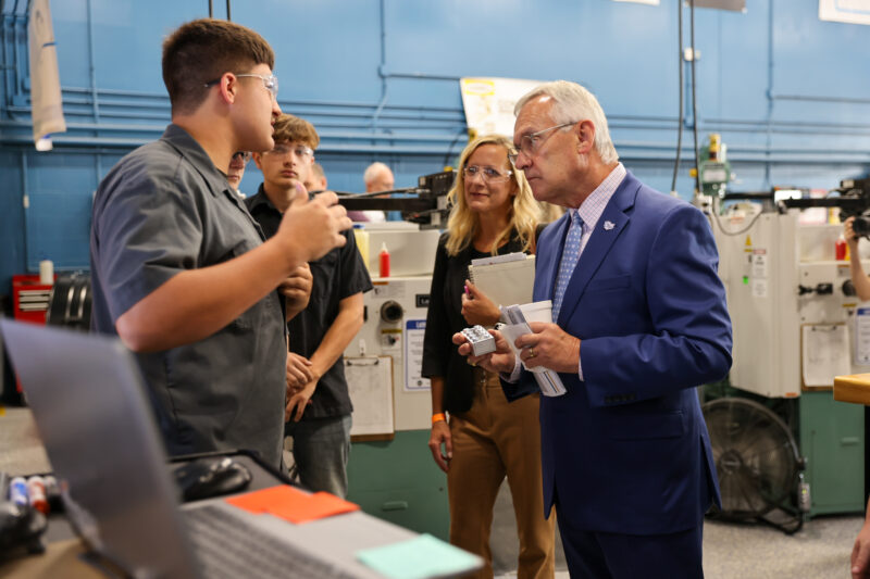 A young man in work attire explains something to an older man in a blue suit—likely from the Business Advisory Council—while two others observe, all inside an industrial workshop at Butler Tech with machinery and blue walls in the background.
