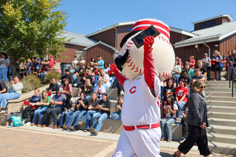 A baseball mascot in a white uniform holds the 2025 Ability Transcends Challenges Award and walks past cheering people seated outdoors on steps and benches during a Project LIFE event on a sunny day.