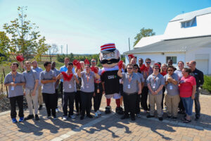 A group of cheerful people in matching gray shirts pose outside on a sunny day with the Project LIFE mascot dressed in a patriotic sailor outfit. With red pom-poms in hand, they celebrate their 2025 Ability Transcends Challenges Award.