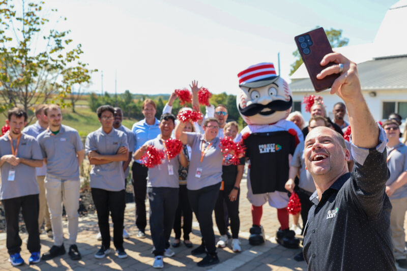 A group of people, including a mascot in a striped hat and workers in gray shirts, pose outside for a cheerful selfie. Celebrating Project LIFE’s 2025 Ability Transcends Challenges Award, they wave red pom-poms and smile under the sunny sky.